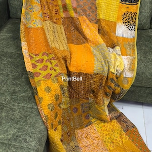 Puede incluir: Una colcha de retazos en tonos amarillos, dorados y marrones, sobre un sofá verde. La colcha presenta una variedad de cuadrados de tela estampada, creando un diseño texturizado y visualmente interesante. Los bordes de la colcha están cuidadosamente acabados.