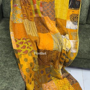 May include: A patchwork quilt in shades of yellow, gold, and brown, draped over a green sofa. The quilt features a variety of patterned fabric squares, creating a textured design. The quilt's edges are neatly finished.