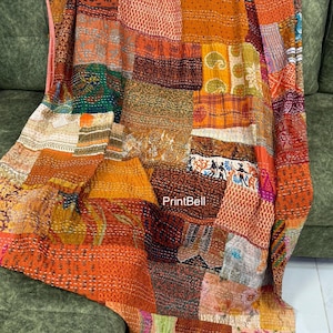 May include: A patchwork quilt with a variety of warm colors, including orange, brown, and red. The quilt is draped over a green sofa. The quilt is made of different fabric squares sewn together, creating a unique and textured design.