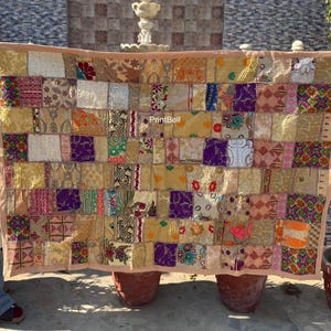 May include: A patchwork textile with a variety of fabrics in shades of gold, purple, pink, and cream. The rectangular piece has a light pink border and is held up by unseen hands. The textile is likely a decorative item.