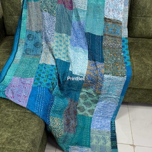 Könnte beinhalten: Eine Patchwork-Decke in Türkis-, Blau- und Lilatönen, drapiert über einem grünen Sofa. Die Decke besteht aus verschiedenen Stoffquadraten, jedes mit einzigartigen Mustern und Texturen. Sichtbare Nähte verleihen dem handgefertigten Textil Details.