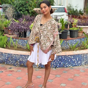 May include: A loose-fitting, olive green kimono-style top featuring a detailed floral pattern in shades of pink and white, with a drawstring waist and long, wide sleeves, paired with a plain white flared skirt and clear heeled sandals.