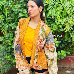 May include: A mustard yellow quilted jacket featuring a vibrant floral pattern with orange, red, and blue petals on a cream background. The jacket has long sleeves, an open front, and a textured finish. The text PrintBell is visible on the right.
