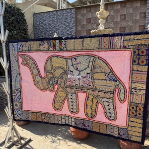 May include: A large, rectangular pink tapestry featuring an embroidered elephant design. The elephant is adorned with gold and blue patterns. The tapestry has a dark blue border with intricate detailing and is supported by two terracotta pots.