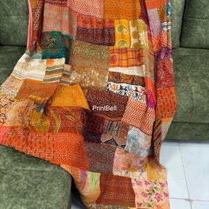 May include: A patchwork quilt with a variety of warm colors, including orange, brown, and red. The quilt is made of different fabric squares with various patterns and textures. The quilt is draped over a green sofa.
