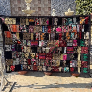 May include: A patchwork textile with a black border and a grid of colourful, patterned squares. The squares feature floral and geometric designs in shades of red, gold, pink, and black. The textile is displayed outdoors.