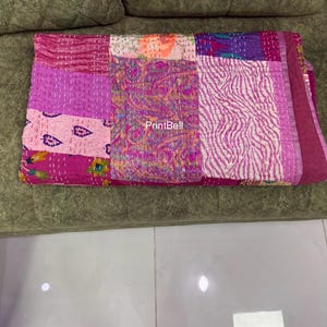 Op de afbeelding: Een gevouwen patchwork quilt in roze, paarse en magenta tinten, met diverse patronen en texturen. De quilt ligt op een groene ondergrond. De tekst "PrintBell" is zichtbaar.