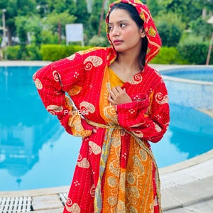 Puede incluir: Una bata con capucha y mangas largas que presenta un diseño de patchwork en rojo y naranja vibrante con intrincados patrones de cachemira color crema y costuras kantha blancas visibles. Incluye un cinturón de tela a juego y el texto PrintBell.