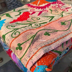 Puede incluir: Una pila de textiles bordados coloridos. El textil superior es de color rosa claro con diseños florales y de vid en rojo, verde y naranja. Los textiles están doblados, revelando capas de colores y patrones vibrantes, incluyendo azul, morado y naranja.