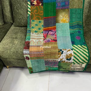 Op de afbeelding: Een patchworkquilt met groene, gele en roze stoffen vierkanten. De quilt is over een groene bank gedrapeerd. De quilt heeft een groene rand en is aan elkaar gestikt met witte draad. De tekst "PrintBell" is zichtbaar op de quilt.
