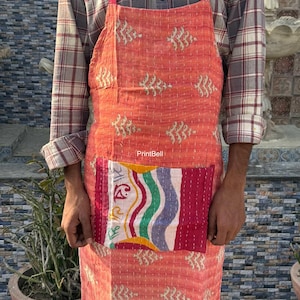 Op de afbeelding: Koraalrood schort met een decoratief patroon en een grote patchwork zak. De zak heeft strepen en andere patronen. Het schort is gemaakt van gewatteerde stof. Afmetingen in centimeters.