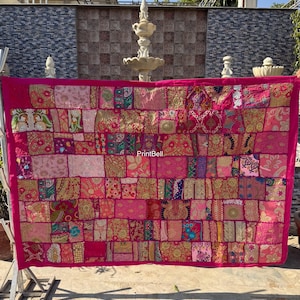 Puede incluir: Un vibrante textil patchwork rosa con una multitud de cuadrados de tela en varios tonos de rosa, rojo y dorado. El textil rectangular, posiblemente un tapiz o una manta, presenta intrincados patrones y texturas en sus numerosos cuadrados.