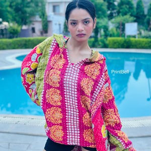 Pode incluir: Um pulôver de manga comprida com capuz, apresentando um design de patchwork vibrante com padrões florais em rosa brilhante, amarelo e verde. A peça tem decote em V, capuz e costuras brancas visíveis. O texto PrintBell é visível.