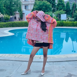 Peut inclure: Un pull à capuche arborant un motif floral multicolore vif dans des tons rose, orange et jaune. Le vêtement présente des manches longues, une coupe ample et le texte PrintBell est visible sur le devant. Il est associé à un short noir.