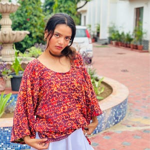 Può includere: Un top dalla vestibilità ampia con un motivo tie-dye bordeaux scuro e arancione con dettagli visibili di cuciture orizzontali bianche, abbinato a una gonna bianca semplice. Il top ha maniche ampie a tre quarti e uno scollo rotondo.