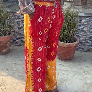 Op de afbeelding: Rood-gele broek met witte ruitvormen en verticale stiksels. De broek heeft een gele tailleband en manchetten. De tekst "PrintBell" is zichtbaar. De persoon draagt een geruit overhemd.