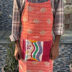 Può includere: Un grembiule color corallo con una tasca patchwork con strisce colorate e testo. Il grembiule ha un motivo a foglie ripetuto e viene indossato con una camicia a quadri. Il grembiule è realizzato in un tessuto morbido e trapuntato.