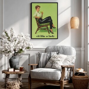 May include: A framed print with a retro illustration of a woman in black leather trousers and heels, holding a cocktail, with the text "sit like a lady" on a green backdrop. The print is in a room with a wooden chair and a vase of white flowers.