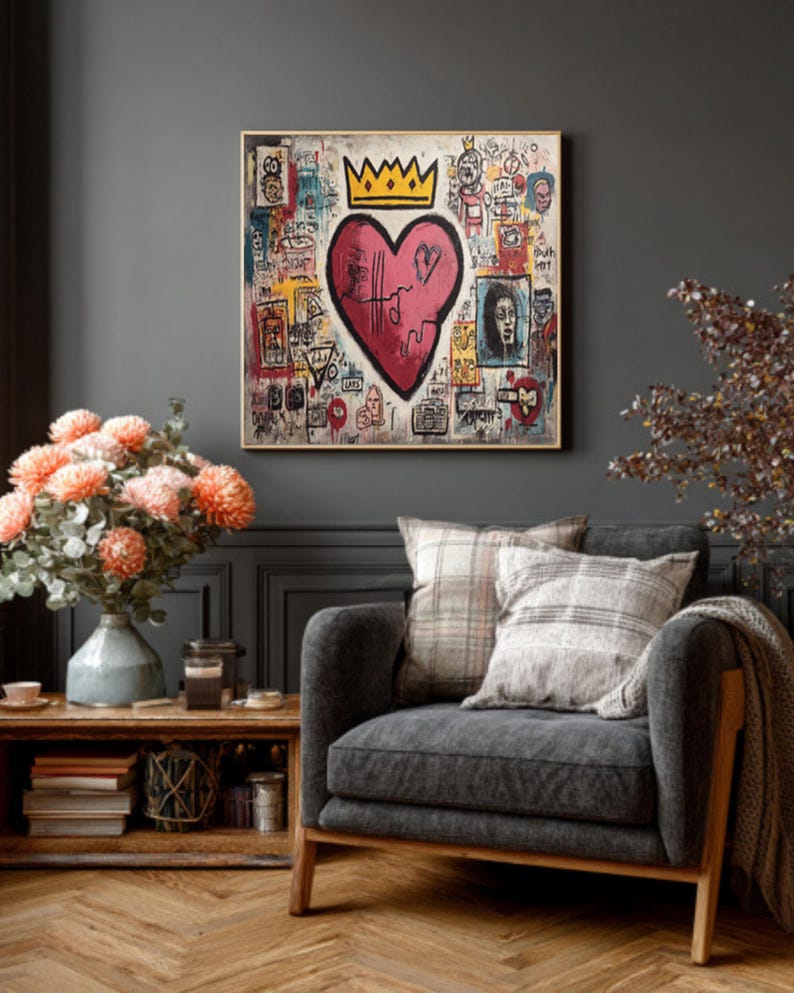 May include: A framed artwork featuring a pink heart with a crown, surrounded by graffiti-style drawings and text. The painting hangs above a gray armchair with plaid pillows, a wooden side table, and a vase of orange flowers.