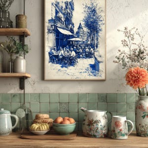May include: A framed blue and white sketch of a European cafe scene. The artwork depicts people seated at tables under an awning, with buildings and trees in the background. The image is displayed above a wooden countertop with decorative pitchers and a bowl of oranges.
