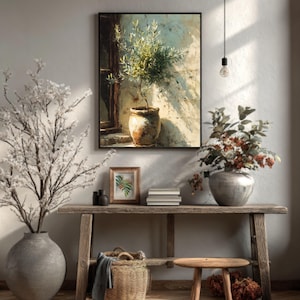 Puede incluir: Pintura enmarcada de un olivo en maceta contra una pared texturizada. Una consola rústica de madera exhibe un jarrón con flores blancas, una impresión botánica enmarcada y una cesta de mimbre. La escena está bañada por la luz natural.