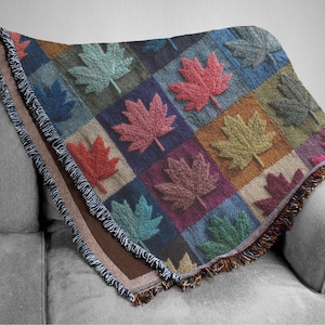 May include: A multi-coloured throw blanket with a repeating pattern of maple leaves in shades of red, pink, green, and blue. The blanket has a fringed edge and is draped over a grey sofa.