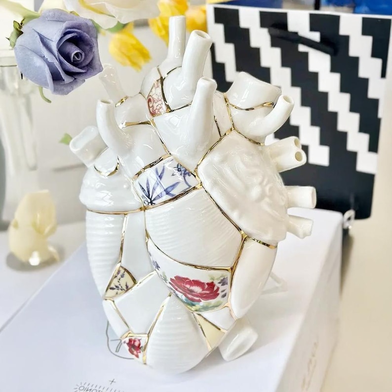 May include: A white ceramic heart-shaped vase with a patchwork design. The vase is adorned with floral patterns and gold accents. The vase has multiple openings and is displayed with flowers and a black and white patterned bag.