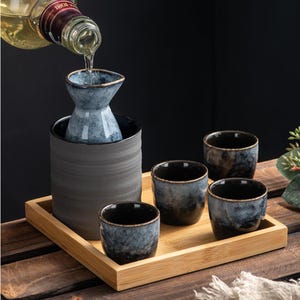 May include: A sake set with a grey ceramic carafe and four matching cups, presented on a bamboo tray. A bottle of sake is pouring into the carafe. The cups and carafe have a blue and black marbled design.