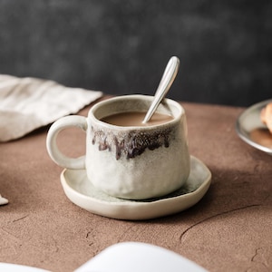 Puede incluir: Una taza de café de cerámica con un platillo a juego. La taza contiene un líquido marrón claro y una cuchara plateada. La taza y el platillo son de color blanquecino con un diseño de goteo marrón alrededor del borde.