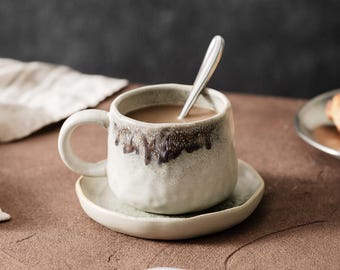 Op de afbeelding: Een keramische koffiekop met bijpassende schotel. De kop is gevuld met een lichtbruine vloeistof en heeft een zilveren lepel. De kop en schotel zijn gebroken wit met een bruin druipontwerp rond de rand.