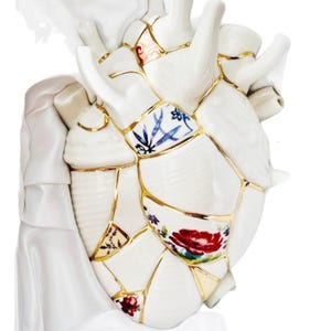May include: A white ceramic heart sculpture, artfully pieced together with gold accents, showcasing floral and patterned designs. The heart's broken sections are meticulously rejoined, creating a unique decorative piece.