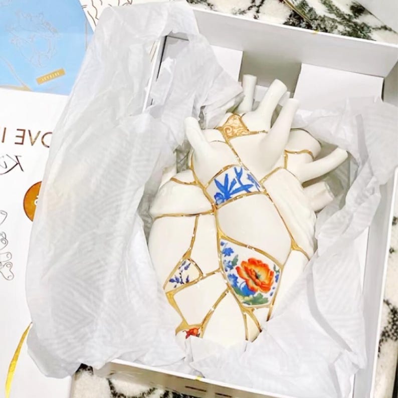 May include: A white ceramic heart sculpture with a kintsugi design, featuring gold accents and floral and geometric patterns. The heart is packaged in a white box with tissue paper.