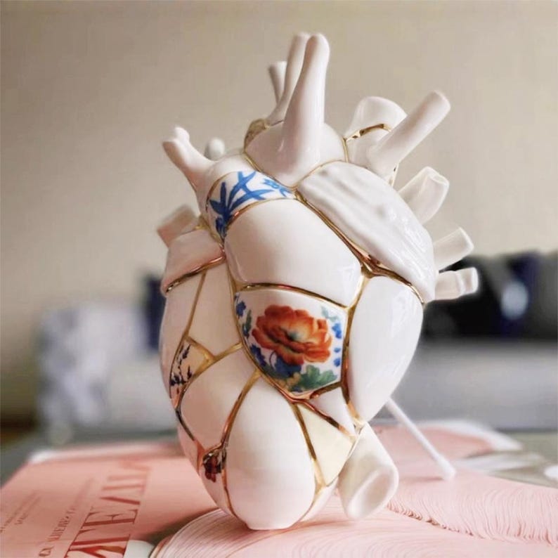 May include: A white ceramic heart sculpture with gold accents and floral patterns. The heart is segmented, with each piece connected by gold lines. Some segments feature blue and white floral designs, while others showcase a red and orange flower.