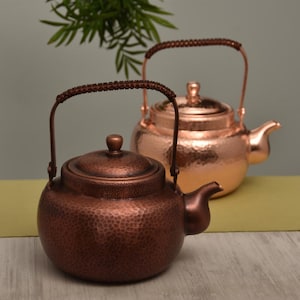 May include: Two copper teapots with hammered textures. The front teapot is a darker copper color, while the one behind it is a lighter copper. Both have rounded bodies, spouts, and handles with woven detailing.
