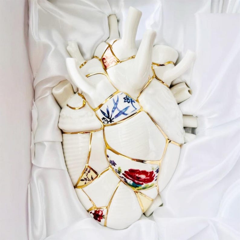 May include: A white ceramic heart sculpture, assembled from broken pieces with gold accents, rests on white satin. The heart features floral and geometric patterns in red, blue, and gold, creating a unique decorative piece.