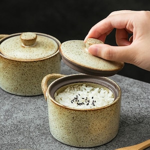 Op de afbeelding: Keramische rijstkom met deksel. De kom is bespikkeld met een lichtbruin glazuur en een donkerbruine rand. Het deksel wordt opgetild om de gekookte rijst binnenin te onthullen. De kom heeft een handvat aan de zijkant.