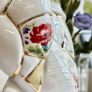 May include: White ceramic vase with a unique design featuring a patchwork of shapes, each outlined in gold. The vase is adorned with red floral patterns and accents of green and purple, creating a striking contrast.