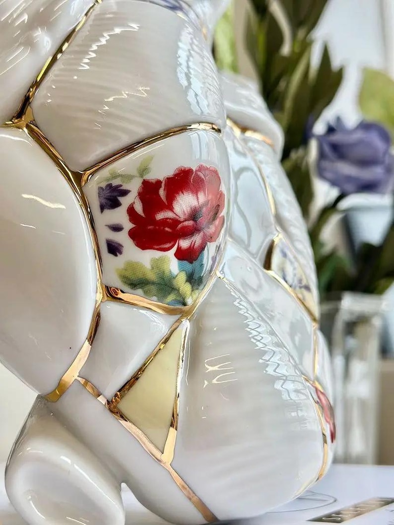 May include: White ceramic vase with a unique design featuring a patchwork of shapes, each outlined in gold. The vase is adorned with red floral patterns and accents of green and purple, creating a striking contrast.
