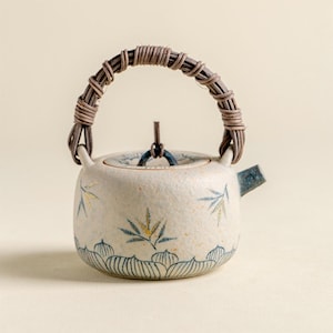 May include: A small, off-white ceramic teapot with a woven handle. The teapot features blue and gold leaf designs and a blue spout. The handle is made of woven brown material. The lid has a small, dark brown handle.