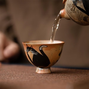 May include: A ceramic teacup with a black cat design is being filled with tea from a matching teapot. The teacup has a tan background with a black cat illustration. The teapot has a brown and black design.