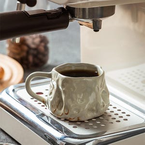 Op de afbeelding: Een lichtgrijze keramische mok met een textuur, organisch ontwerp, gevuld met donkere koffie. De mok staat op een roestvrijstalen lekbak van een espressomachine, met de componenten van de machine zichtbaar op de achtergrond. Koffiebonen zijn zichtbaar in een glazen pot.