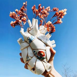 May include: A white ceramic heart-shaped vase with a kintsugi design, featuring gold accents and floral patterns. The vase holds dried orange flowers against a clear blue sky. The vase is held by a person's hand.
