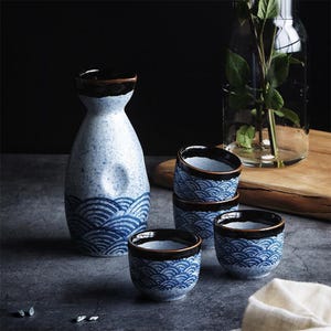 May include: A ceramic sake set featuring a blue and white wave pattern. The set includes a tall, slender bottle and several small cups. The rims of the bottle and cups are dark brown. The set is on a dark gray surface.