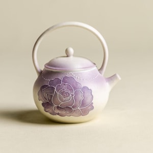 May include: A ceramic teapot with a white and purple gradient finish. A large purple rose design is painted on the side. The teapot has a curved handle and a small spout.