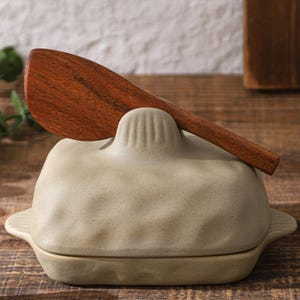 Peut inclure: Un beurrier en céramique beige avec un couvercle assorti et une cuillère en bois. Le plat a une surface texturée et de petites poignées. La cuillère en bois repose sur le couvercle. Mesure environ 15 cm de long.