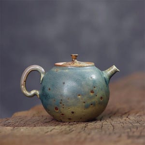 Puede incluir: Una pequeña tetera redonda de cerámica de color verde azulado con tapa a juego. La tetera tiene un asa curva y un pico. La superficie tiene un patrón moteado con manchas marrones. La tetera está sobre una superficie de madera.