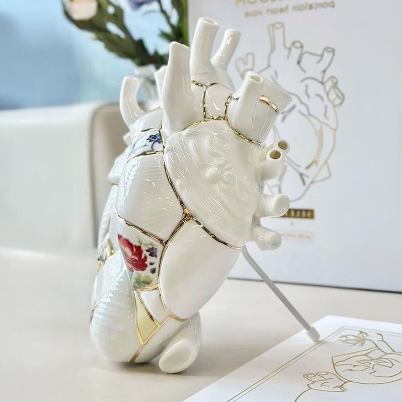 May include: A white porcelain heart sculpture with gold accents, featuring a kintsugi design. The heart is decorated with floral patterns and has a unique, artistic appearance. A card with a heart illustration is in the background.