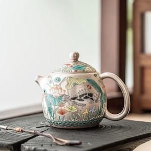 Könnte beinhalten: Eine silberne Teekanne mit Deckel, Henkel und Ausguss. Die Teekanne ist mit einem farbenfrohen Design aus Fischen, Lotusblumen und anderen Wasserpflanzen verziert. Die Teekanne steht auf einer dunklen Holzoberfläche.