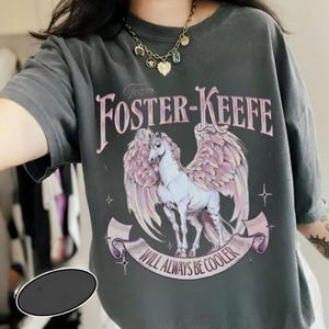 Può includere: T-shirt grigio scuro con una grafica di cavallo alato e il testo "Team Foster-Keefe" sopra e "Will Always Be Cooler" sotto. Il design include accenti rosa e bianchi.