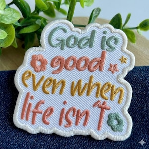 May include: Embroidered patch with the text "God is good even when life isn't". The patch features a white background with colorful lettering in green, peach, and gold. Decorative floral and star accents are included.